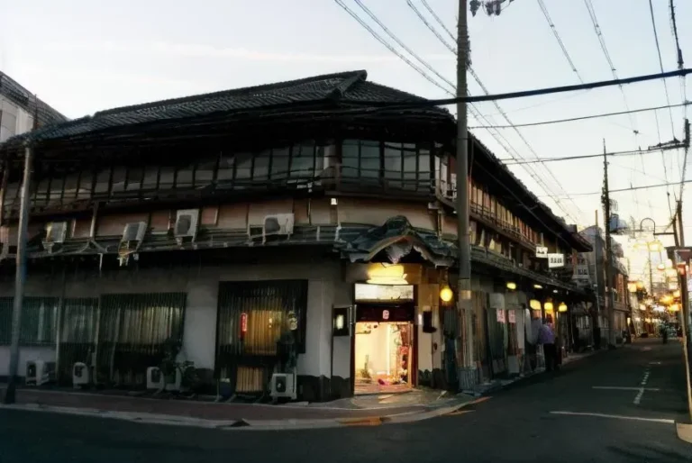 日本の街角にある建物を日没時に描いた写実的な画像。複雑なディテールとリアルなライティングが特徴。